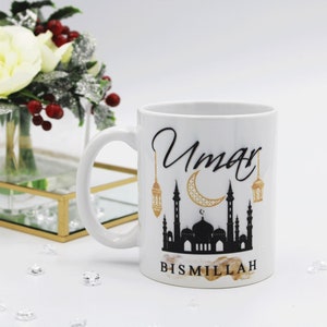 Pode incluir: Caneca de cerâmica branca com um desenho de mesquita em preto e dourado, o nome "Umar" em escrita cursiva e a palavra "Bismillah". A caneca tem uma alça e é decorada com uma lua crescente e lanternas.