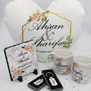 May include: A wedding gift set featuring a white heart-shaped cushion with gold accents and the names "Ahsan & Sharifa" printed on it. The set also includes a slate plaque with the same names and a floral design, two white mugs with the names and a floral design, and two keychains with the names and a floral design.