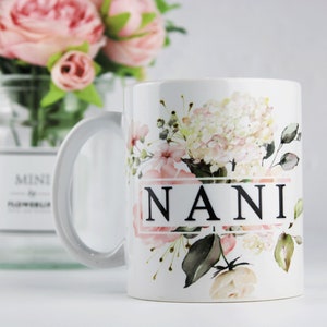 Pode incluir: Caneca de cerâmica branca com um design floral com flores rosa e brancas. A caneca tem uma alça preta e a palavra "NANI" impressa em preto na frente.