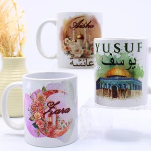 Pode incluir: Três canecas de cerâmica branca com designs personalizados. Uma caneca apresenta uma lua crescente floral com o nome "Zara". Outra exibe uma ilustração de uma mesquita com o nome "Aisha". A terceira caneca tem uma ilustração de cúpula com o nome "Yusuf".