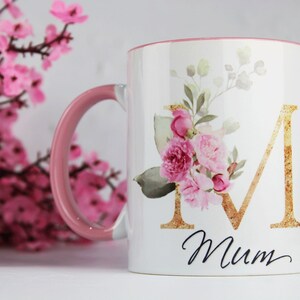 Pode incluir: Caneca de cerâmica branca com borda e alça rosa. A caneca apresenta uma letra dourada "M" cercada por flores rosa em aquarela e folhas verdes. A palavra "Mum" está escrita em itálico preto abaixo da letra.