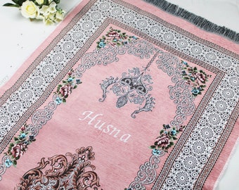 May include: A pink prayer rug with a white and silver floral design. The rug has a fringe border and the name "Husna" is embroidered in the center.