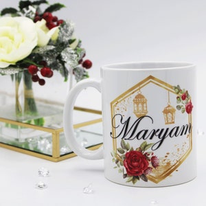 Pode incluir: Caneca de cerâmica branca com um design hexagonal dourado com um padrão floral, duas lanternas e o nome "Maryam" em escrita preta.