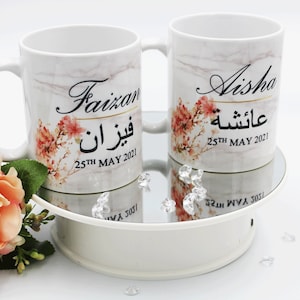Pode incluir: Duas canecas de cerâmica branca com designs de mármore. As canecas apresentam detalhes florais e nomes personalizados, "Faizan" e "Aisha", com a data "25 de MAIO de 2021" impressa abaixo de cada nome.