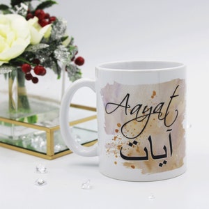 Pode incluir: Caneca de cerâmica branca com um design em aquarela em tons de marrom e rosa. A caneca apresenta o nome "Aayat" em caligrafia preta e a palavra árabe para "você" em letras douradas.