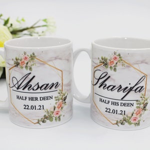 Pode incluir: Duas canecas de cerâmica branca com detalhes dourados e desenhos florais. As canecas têm os nomes "Ahsan" e "Sharija" impressos, juntamente com o texto "Half her Deen" e "Half his Deen" e a data "22.01.21".