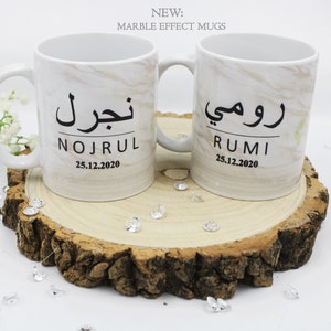 Pode incluir: Duas canecas de cerâmica branca com efeito mármore, cada uma personalizada com um nome e data em letras pretas. As canecas estão sobre uma fatia de madeira, rodeadas por cristais e flores decorativas. O texto "NEW: MARBLE EFFECT MUGS" está no topo.