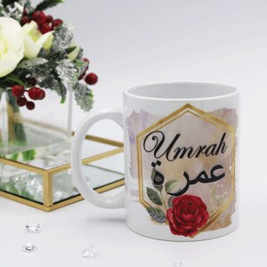 Pode incluir: Caneca de café de cerâmica branca com um design hexagonal dourado com as palavras "Umrah" e caligrafia árabe. O design também inclui uma rosa vermelha com folhas verdes.