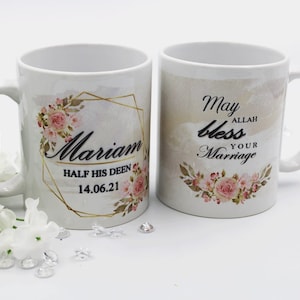 Pode incluir: Duas canecas de cerâmica branca com desenhos florais e detalhes dourados. Uma caneca diz "Mariam Half His Deen 14.06.21" e a outra caneca diz "Que Allah abençoe seu casamento".