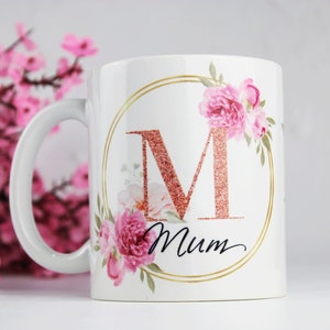 Pode incluir: Caneca de cerâmica branca com uma borda dourada e um design floral. O design apresenta uma letra "M" de glitter rosa em um círculo com flores rosas e folhas verdes. A palavra "Mum" está escrita em itálico preto abaixo da letra.