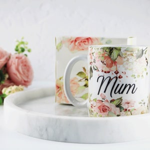 Pode incluir: Caneca de cerâmica branca com um design floral e a palavra "Mum" impressa em preto. A caneca está sobre uma bandeja de mármore branco.