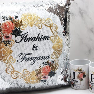Pode incluir: Uma almofada de lantejoulas prateadas com um design floral branco e os nomes "Ibrahim & Farzana" em letras pretas. Também são mostradas duas canecas brancas com desenhos florais e as iniciais "I" e "F".