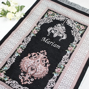 May include: Black prayer rug with a white and silver floral border. The rug has a pink and silver design in the centre and the name "Mariam" is embroidered in the centre.