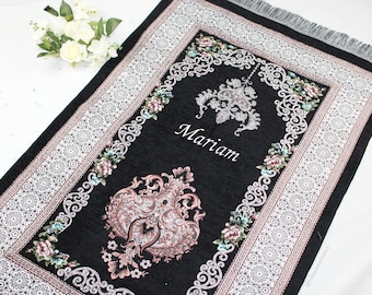 May include: Black prayer rug with a white and silver floral border. The rug has a pink and silver design in the center and the name "Mariam" is embroidered in the center.