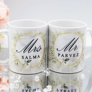 Pode incluir: Duas canecas de cerâmica branca com detalhes dourados e desenhos florais em aquarela verde. As canecas têm o texto "Mrs. Salma" e "Mr. Parvez" impresso nelas.
