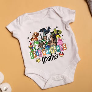 May include: White baby bodysuit with a colorful graphic featuring cartoon characters and the text "EPIC UNIVERSE Brother". The design includes a castle, stars, and a mushroom.