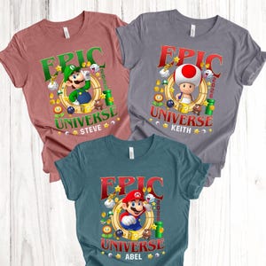 May include: Three short-sleeve t-shirts in mauve, gray, and teal. Each features a graphic with the text EPIC UNIVERSE 2026, a circular frame, and Nintendo characters: Luigi with the name STEVE, Toad with KEITH, and Mario with ABEL.