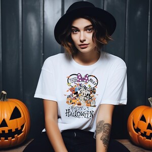 May include: White t-shirt with a Halloween-themed graphic featuring cartoon characters, a purple bow, and the name "Riley." The shirt also has the words "Happy Halloween" printed on it. Two carved pumpkins flank the person wearing the shirt.