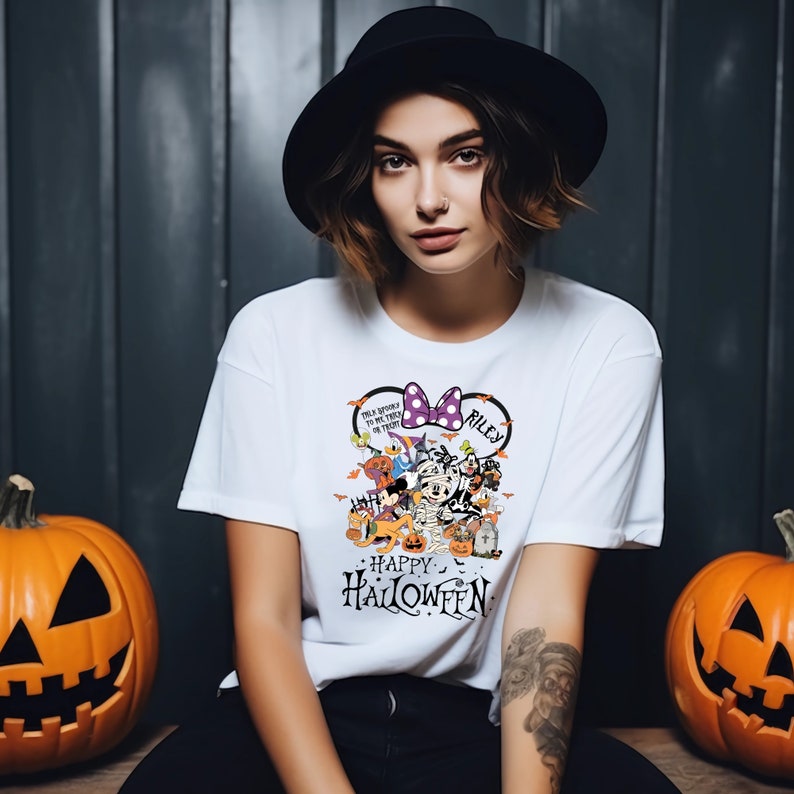 May include: White t-shirt with a Halloween-themed graphic featuring cartoon characters, a purple bow, and the name "Riley." The shirt also has the words "Happy Halloween" printed on it. Two carved pumpkins flank the person wearing the shirt.