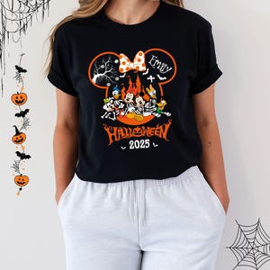 以下が含まれることがあります： ハロウィーンをテーマにしたデザインの黒いTシャツ。デザインには、水玉模様のリボンが付いたミッキーマウスの耳、「Emily」という名前、スケルトンのコスチュームを着たディズニーのキャラクターが含まれています。「Halloween 2025」というテキストも表示されています。