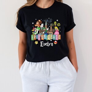 May include: Black t-shirt featuring a colorful graphic with the text "EPIC UNIVERSE" and the name "Evelyn". The graphic includes cartoon characters from various franchises, such as Donkey Kong, Mario, and Princess Peach.