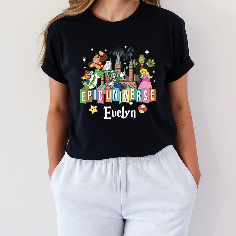 May include: Black t-shirt featuring a colorful graphic with the text "EPIC UNIVERSE" and the name "Evelyn". The graphic includes cartoon characters from various franchises, such as Donkey Kong, Mario, and Princess Peach.