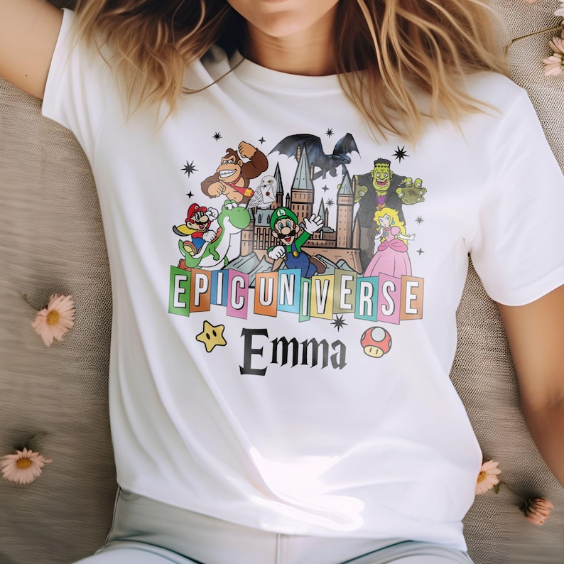 May include: White t-shirt featuring a colorful graphic with cartoon characters, the text "EPIC UNIVERSE" and the name "Emma". The design includes a castle, a dragon, and various video game characters.