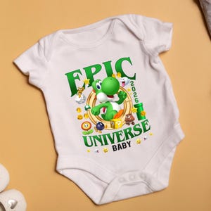 May include: A white short-sleeve baby bodysuit featuring a graphic print of a green Yoshi character inside a gold ring. The text reads EPIC UNIVERSE 2026 BABY, surrounded by colorful game icons including coins, a question block, a pipe, and stars.