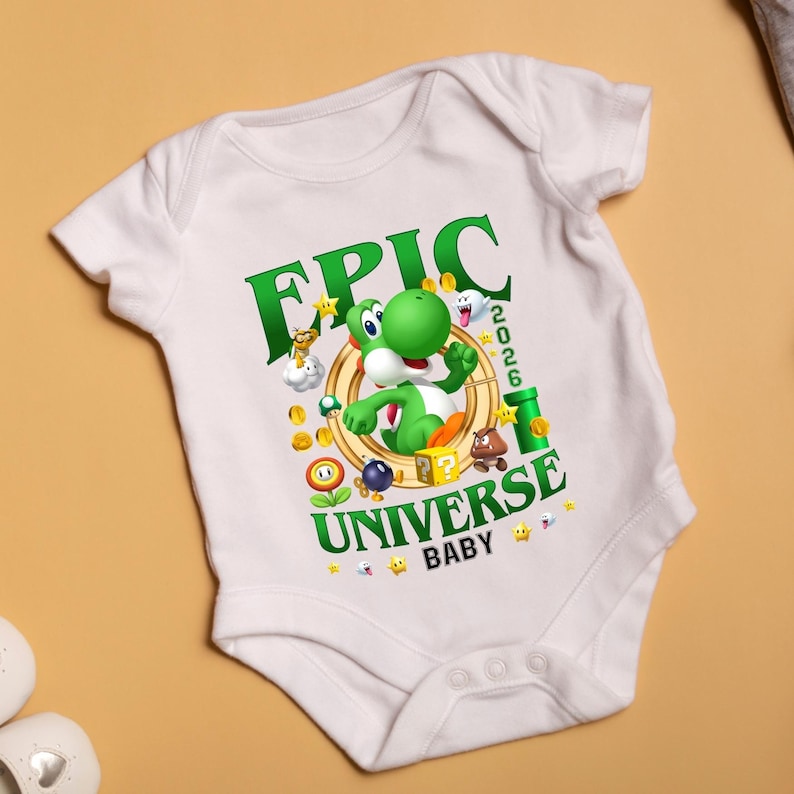 May include: A white short-sleeve baby bodysuit featuring a graphic print of a green Yoshi character inside a gold ring. The text reads EPIC UNIVERSE 2026 BABY, surrounded by colorful game icons including coins, a question block, a pipe, and stars.