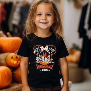 以下が含まれることがあります： ディズニーキャラクターが描かれたハロウィーンをテーマにした黒いTシャツ。デザインには、リボンが付いたミニーマウスのシルエット、「Emily」という名前、そして「Halloween 2025」という文字が含まれています。このTシャツは小さな子供が着ています。