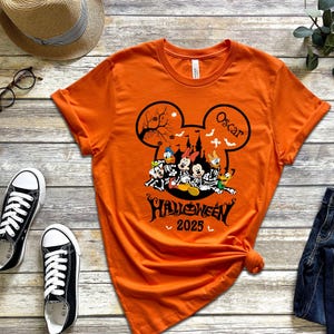 以下が含まれることがあります： ハロウィーンのデザインが施されたオレンジ色のTシャツ。デザインにはミッキーマウスの耳、不気味なお城、漫画のキャラクターが含まれています。シャツのテキストは「ハロウィーン2025」と「オスカー」という名前です。