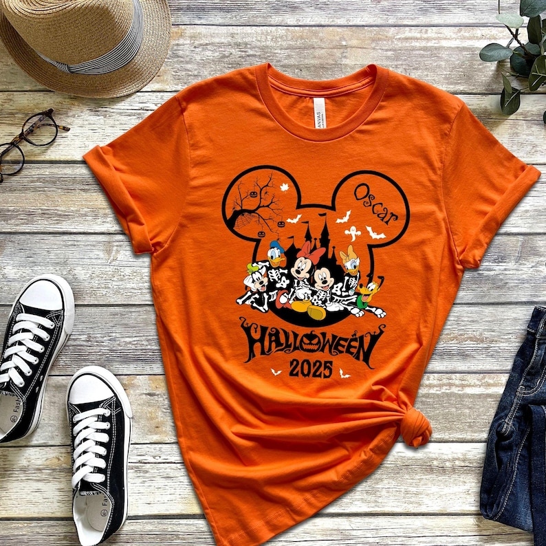 以下が含まれることがあります： ハロウィーンのデザインが施されたオレンジ色のTシャツ。デザインにはミッキーマウスの耳、不気味なお城、漫画のキャラクターが含まれています。シャツのテキストは「ハロウィーン2025」と「オスカー」という名前です。