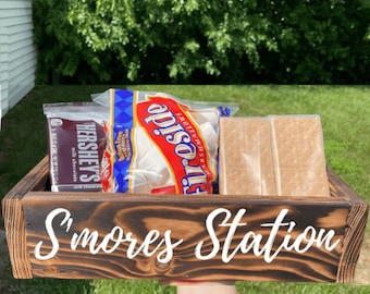 May include: A wooden box with the words "S'mores Station" painted on the front. The box is filled with graham crackers, marshmallows, and chocolate bars.