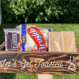 May include: A wooden s'mores kit with Hershey's chocolate, marshmallows, and graham crackers. The kit is in a dark brown wooden tray with the words "Let's Get Toasted" and a campfire graphic.