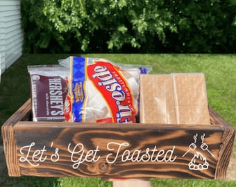May include: A wooden s'mores kit with Hershey's chocolate, marshmallows, and graham crackers. The kit is in a dark brown wooden tray with the words "Let's Get Toasted" and a campfire graphic.