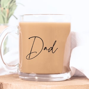 Peut inclure: Une tasse en verre transparent remplie d'un liquide brun clair, peut-&ecirc;tre du caf&eacute; ou du th&eacute;. Le mot "Dad" est &eacute;crit en script noir &eacute;l&eacute;gant sur le devant. La tasse est pos&eacute;e sur une surface en bois.