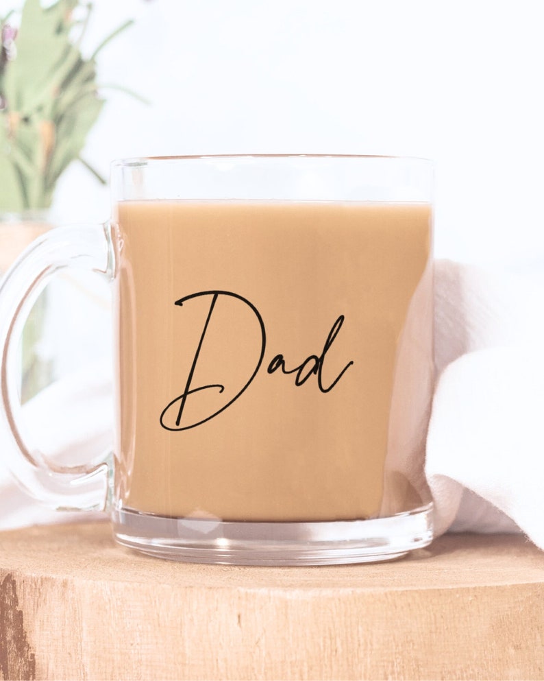 Peut inclure: Une tasse en verre transparent remplie d'un liquide brun clair, peut-&ecirc;tre du caf&eacute; ou du th&eacute;. Le mot "Dad" est &eacute;crit en script noir &eacute;l&eacute;gant sur le devant. La tasse est pos&eacute;e sur une surface en bois.