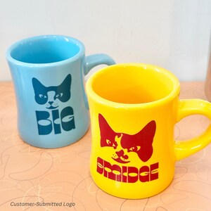 以下が含まれることがあります： 猫の顔のグラフィックが描かれた、水色と黄色の2つのセラミックマグ。水色のマグには猫の顔の下に「BIG」の文字。黄色のマグには猫の顔の下に「BRIDGE」の文字。どちらのマグもカーブしたハンドルが付いています。