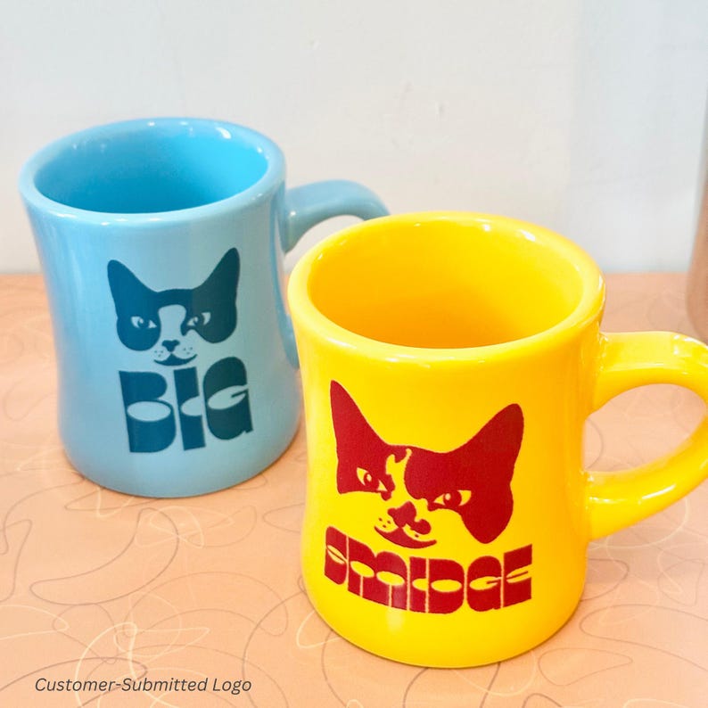 以下が含まれることがあります： 猫の顔のグラフィックが描かれた、水色と黄色の2つのセラミックマグ。水色のマグには猫の顔の下に「BIG」の文字。黄色のマグには猫の顔の下に「BRIDGE」の文字。どちらのマグもカーブしたハンドルが付いています。