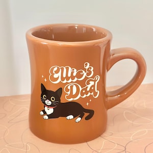 以下が含まれることがあります： カーブしたハンドルが付いた茶色のセラミックマグ。マグには、白い文字で「Ellie's Dad」と、白い足と赤い首輪をした黒猫の漫画イラストが描かれています。マグは広い開口部を持っています。