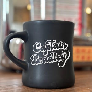 May include: A black ceramic mug with a curved handle. The mug features the white text "Captain Bradley" in a retro font. The mug is sitting on a wooden surface.