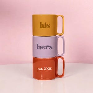 May include: Three stacked ceramic mugs in mustard yellow, lavender, and rust orange. The top mug says "his", the middle mug says "hers", and the bottom mug says "est. 2026". Each mug has a curved handle.