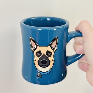 以下が含まれることがあります： 犬のイラストが描かれた青いセラミックマグカップ。犬はベージュと黒の模様、茶色の目、青い首輪とタグが付いています。マグカップは湾曲したハンドルと光沢のある仕上げが特徴です。