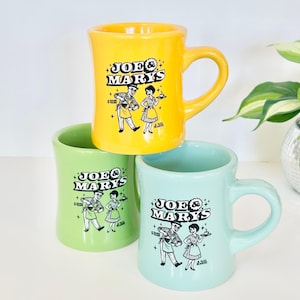 May include: Three ceramic mugs in yellow, light green, and pale blue. Each mug features a black graphic print of a man and woman with the text "JOE & MARY'S" and "A DARK O'BARK" below. The mugs have a sturdy handle and a smooth, glossy finish.
