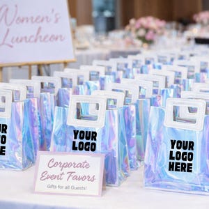 May include: Iridescent tote bags with white handles and the text "YOUR LOGO HERE" are arranged on a white surface. A sign reads "Corporate Event Favors" and "Gifts for all Guests!" A sign in the background reads "Women's Luncheon".