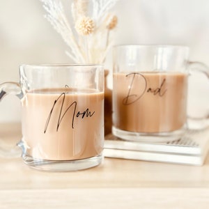 Peut inclure: Deux tasses en verre transparent remplies d'un liquide brun clair, probablement du caf&eacute; ou du th&eacute;. La tasse de gauche porte le mot "Mom" en &eacute;criture noire, et celle de droite "Dad". Les deux tasses ont des anses.