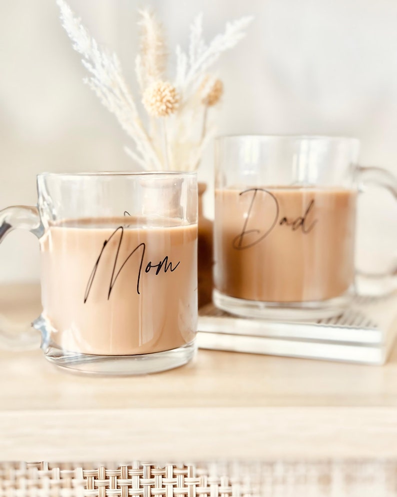 Peut inclure: Deux tasses en verre transparent remplies d'un liquide brun clair, probablement du caf&eacute; ou du th&eacute;. La tasse de gauche porte le mot "Mom" en &eacute;criture noire, et celle de droite "Dad". Les deux tasses ont des anses.