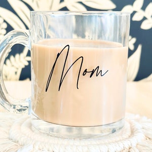 Peut inclure: Tasse en verre transparent remplie d'une boisson brun clair, avec le mot "Mom" &eacute;crit en noir. La tasse est pos&eacute;e sur un dessous de verre en macram&eacute; blanc. Le fond pr&eacute;sente un motif floral bleu fonc&eacute; et blanc.