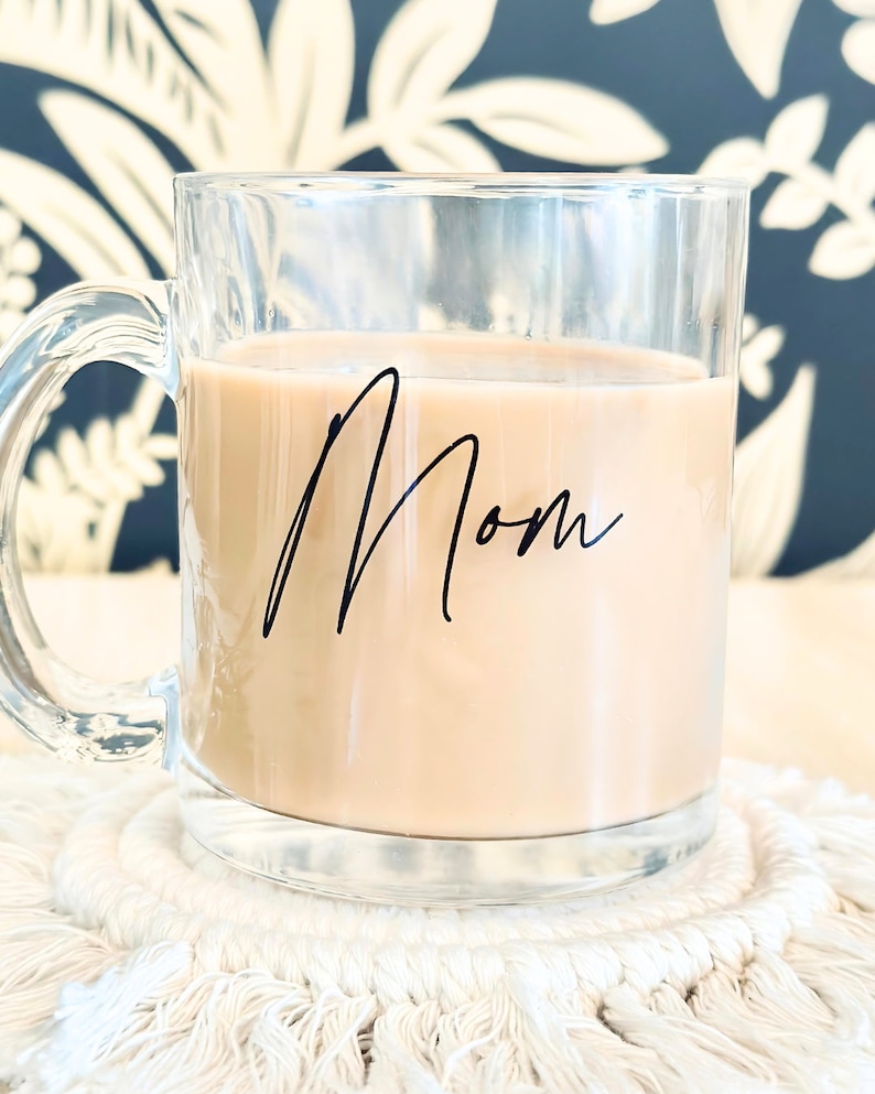 Peut inclure: Tasse en verre transparent remplie d'une boisson brun clair, avec le mot "Mom" &eacute;crit en noir. La tasse est pos&eacute;e sur un dessous de verre en macram&eacute; blanc. Le fond pr&eacute;sente un motif floral bleu fonc&eacute; et blanc.