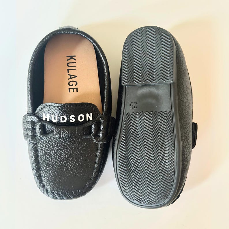 May include: Black leather loafers with the name "HUDSON" across the top. The inside of the shoe has the name "KULAGE" printed on it. The sole of the shoe is black with a textured pattern. The size is 25.